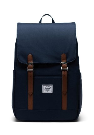 Navy blue backpack made of fabric with brown leather straps and metallic buckles. Features a front pocket and logo patch.