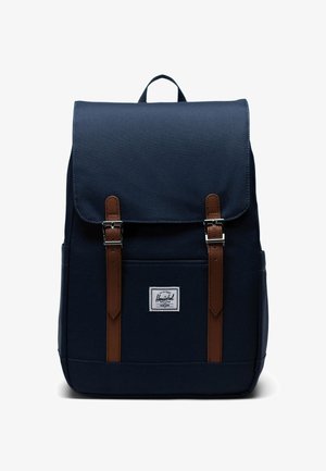 Sac à dos bleu marine en tissu avec des sangles en cuir brun et des boucles métalliques. Il comporte une poche avant et un patch logo.