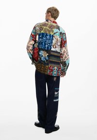Camisa de patchwork multicolor con varios patrones florales y abstractos; corte holgado. Combinada con jeans azul oscuro con parches decorativos.