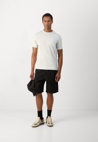 Lichtgrijs T-shirt met korte mouwen, gecombineerd met zwarte shorts, en groene en beige sneakers. Houdt een zwarte pet vast en staat op een witte achtergrond.