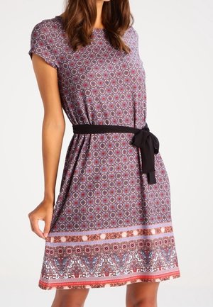 Vrouw draagt een jurk met korte mouwen, versierd met bloem- en geometrische patronen, afgewerkt met een zwarte stoffen ceintuur die in de taille is gestrikt.