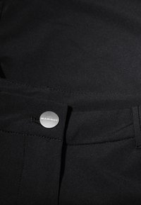 Černé kalhoty s elegantním designem, s stříbrným knoflíkem označeným "MAMMUT", hladkou texturou a minimalistickým prošíváním podél pásku.