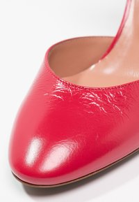 Chaussure à talons hauts rouge avec une texture lisse et brillante, un bout pointu et un dos arrondi. Matériau en cuir avec des accents de couture visibles.