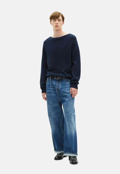 Dunkelblauer Strickpullover mit weitem Ausschnitt, kombiniert mit hellblauen weiten Jeans und schwarzen Lederschuhen.