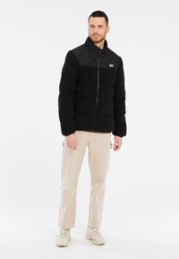 Sort fleece pufferjakke med en skinnende sort øverste sektion, fuld lynlås, stående krave og elastiske manchetter, båret over beige bukser og sko.