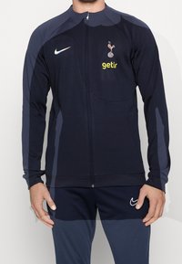 Giacca a zip atletica navy realizzata in tessuto liscio, presenta un logo e una stampa dello sponsor sul petto, con maniche lunghe e vestibilità slim.