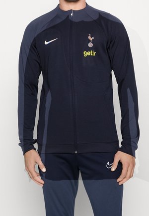 Marinefarbener sportlicher Reißverschlussjacke aus glattem Stoff, mit einem Logo und Sponsor-Druck auf der Brust, langen Ärmeln und einer schlanken Passform.