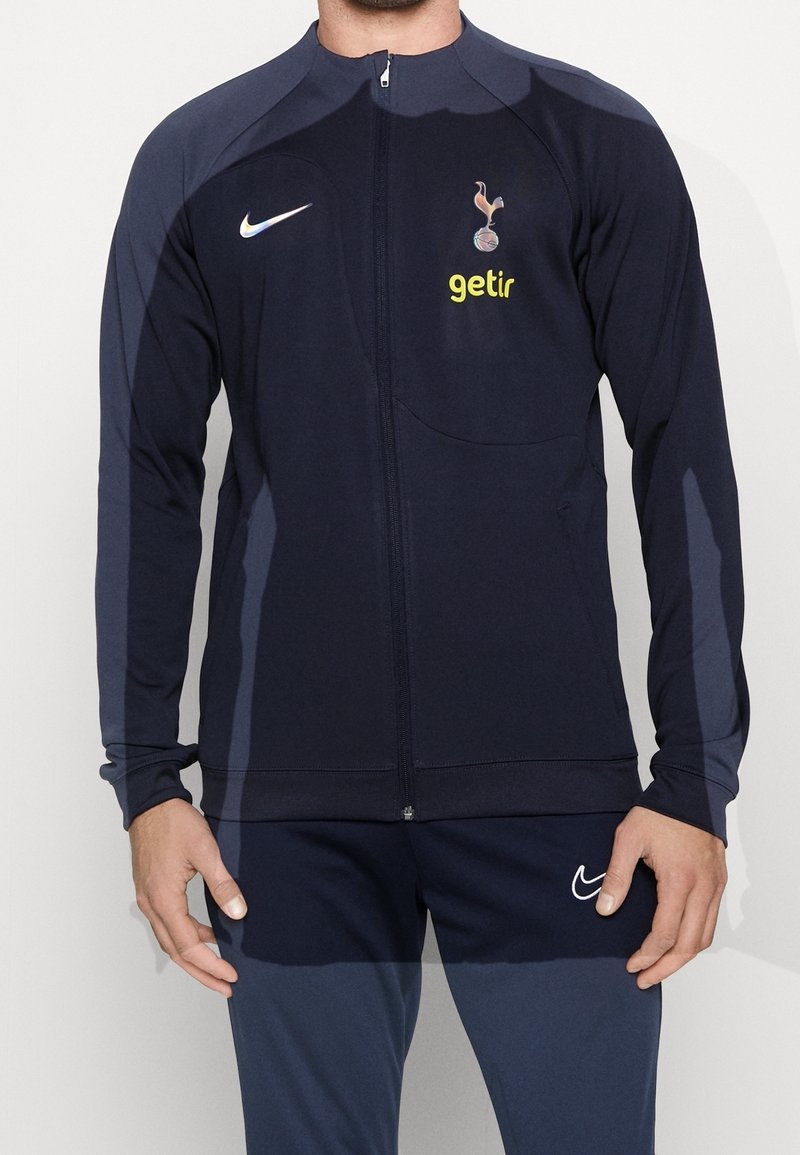 Giacca a zip atletica navy realizzata in tessuto liscio, presenta un logo e una stampa dello sponsor sul petto, con maniche lunghe e vestibilità slim.