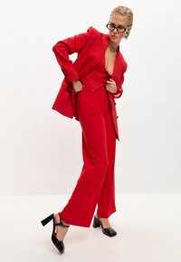 Blazer rosso su misura con pantaloni abbinati, caratterizzato da linee nette, un profondo scollo e scarpe nere con tacco a blocco. Tessuto morbido e design aderente.