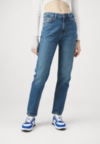 Vaqueros de mezclilla azul con corte recto, cintura alta y un sutil detalle de desteñido en la parte frontal. Combinados con zapatillas blancas y azules.