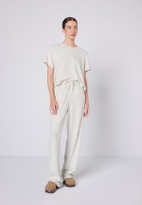 Žena stojící, oblečená v světlém béžovém tričku volného střihu a odpovídajících kalhotách se stahovací šňůrkou, na nohou má hnědé dřeváky, na neutrálním šedém pozadí.