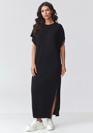 Kobieta o długich ciemnych włosach ubrana w czarną maxi sukienkę z krótkim rękawem i bocznym rozcięciem oraz białe sneakersy na platformie, stojąca na jasnym tle.