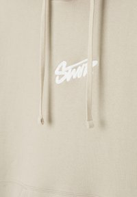 Beige Kapuzenpullover mit einem weißen gestickten Logo. Er hat eine Kordelzugkapuze und einen weichen, strukturierten Stoff sowie eine Kängurutasche.