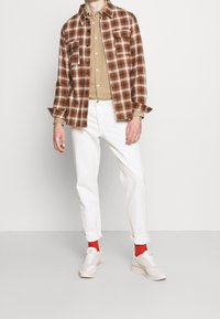 Karierte braun-weiß-karierte Flanellhemd über einem beigen Hemd, kombiniert mit weißen Jeans und roten Socken, abgerundet mit weißen Turnschuhen.