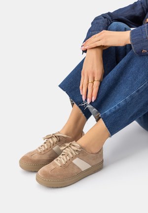 Osoba nosící béžové semišové tenisky s bílým pruhem, modré džíny s roztřepeným lemem a zlaté prsteny na prstech, sedící s rukama opřenýma o kolena.