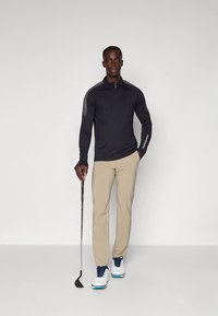 Langärmliges schwarzes Golfshirt mit Reißverschluss am Kragen und strukturierten Schulterpaneelen, kombiniert mit beigen Hosen und weißen Golfschuhen mit blauen Akzenten.