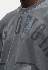 Γκρι βαμβακερό φούτερ με μεγάλες, ξεθωριασμένες μαύρες γραφικές αποτυπώσεις και μικρή μπλε λεπτομέρεια. Ριμπ λαιμόκοψη και μαλακό, υφασμάτινο ύφασμα με υφή.