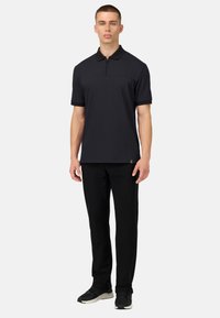 Camisa polo negra de manga corta con cuello con cremallera, combinada con pantalones negros y zapatillas oscuras. Tejido suave, diseño ajustado.