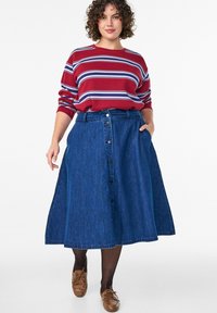 Džínsová áčková sukňa tmavomodrej farby s predným zapínaním na gombíky, bočnými vreckami a lemom dosahujúcim pod kolená. Kombinovaná s červeným pruhovaným svetríkom.