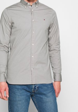 Mężczyzna ubrany w jasnoszarą koszulę z długim rękawem zapinaną na guziki z małym logo na piersi oraz niebieskie dżinsy, stojący na jednolitym tle.