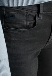 Los jeans de mezclilla negra presentan un ajuste slim con un lavado oscuro, bolsillo frontal y un acento de logo azul. La cinturilla es ligeramente visible con un su éter azul claro.