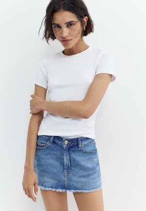 Jeune femme aux cheveux courts et foncés portant un t-shirt blanc et une mini-jupe en jean bleue, debout devant un fond blanc.