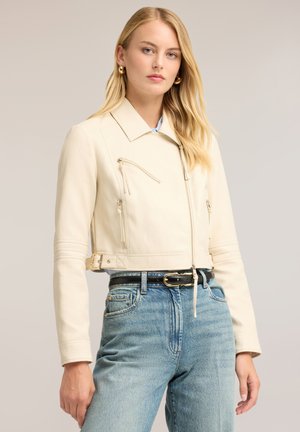 Lyshåret kvinde iført en cremefarvet cropped jakke med lynlåsdetailler, guldringeøreringe, sort bælte og lyseblå højtaljede jeans.