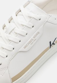 Biały skórzany sneaker z materiałowymi sznurowadłami, z beżowym paskiem akcentującym i wytłoczonym logo. Gładka tekstura z niskim fasonem.