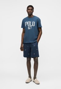 Mann trägt dunkelblaues "POLO 67" T-Shirt und gemusterte dunkelblaue Shorts, steht mit einer Hand in der Tasche und beigen Loafern.
