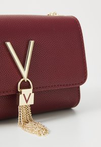 Burgundy textured leather bag with a gold V logo accent and a gold chain tassel detail, featuring a structured rectangular shape.