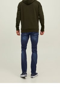 Olivgrön hoodie med en bakficka, blå jeans med slim fit och mörka skor. Stofferna ser släta ut med en avslappnad design.