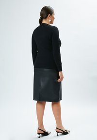 Schwarzer, figurbetonter Pullover mit langen Ärmeln, kombiniert mit einem schwarzen Leder-Bleistiftrock. Das Modell trägt schwarze Slingback-Absätze mit Verzierungen.