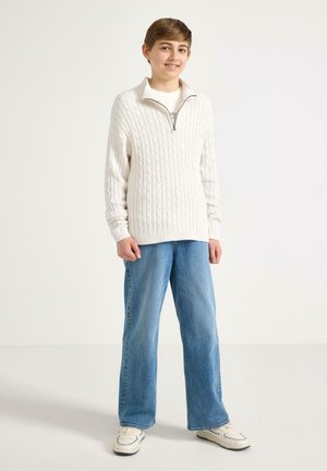 Dreng stående og smilende, iført hvid kabelstrikket sweater med lynlås, lyseblå jeans og hvide sneakers, mod en ensfarvet hvid baggrund.