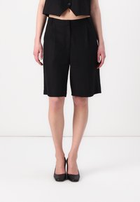 Svarta skräddarsydda shorts i lätt tyg, med slät framsida, sidofickor och knälång passform, tillsammans med svarta högklackade skor.