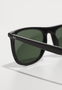 Ochelari de soare din plastic negru cu lentile verzi, având o formă rectangulară elegantă și un logo argintiu pe ramă. Textură netedă cu finisaj lustruit.