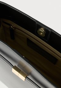 Schwarze Lederhandtasche mit glänzendem Finish, goldfarbenem Verschluss und beigem Stofffutter mit Reißverschlusstasche. Offenes Oberteil.