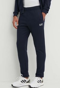 Pantaloni della tuta blu navy realizzati in tessuto morbido, con vita elastica, gambe affusolate e un piccolo logo EA7 sulla coscia superiore.