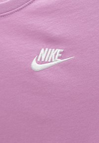 Λευκό λογότυπο Nike κεντημένο σε ροζ ύφασμα, εμφανίζοντας το όνομα της μάρκας και το σύμβολο swoosh σε υλικό ρουχισμού.