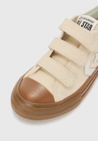 Beige lerret sneakers med tre krok- og løkkestropp, brun gummitåhette og en teksturert såle med fiskebensmønster.