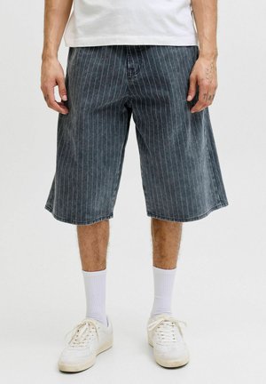 Mand iført sorte knælang shorts med striber, hvide crew-sokker og hvide sneakers, stående mod en ensfarvet baggrund.