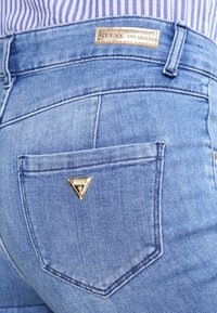 Jeans de mezclilla de color azul claro con parche del logo de Guess encima del bolsillo trasero y un pequeño emblema triangular dorado de la marca en el bolsillo.