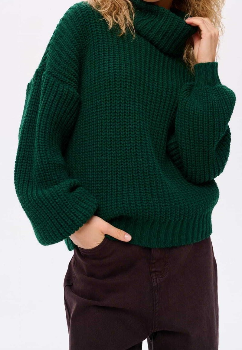 Grøn strikket sweater med høj krave, afslappet pasform og sænkede skuldre. Ribbet manchetter og kant. Sammen med mørkebrune bukser.