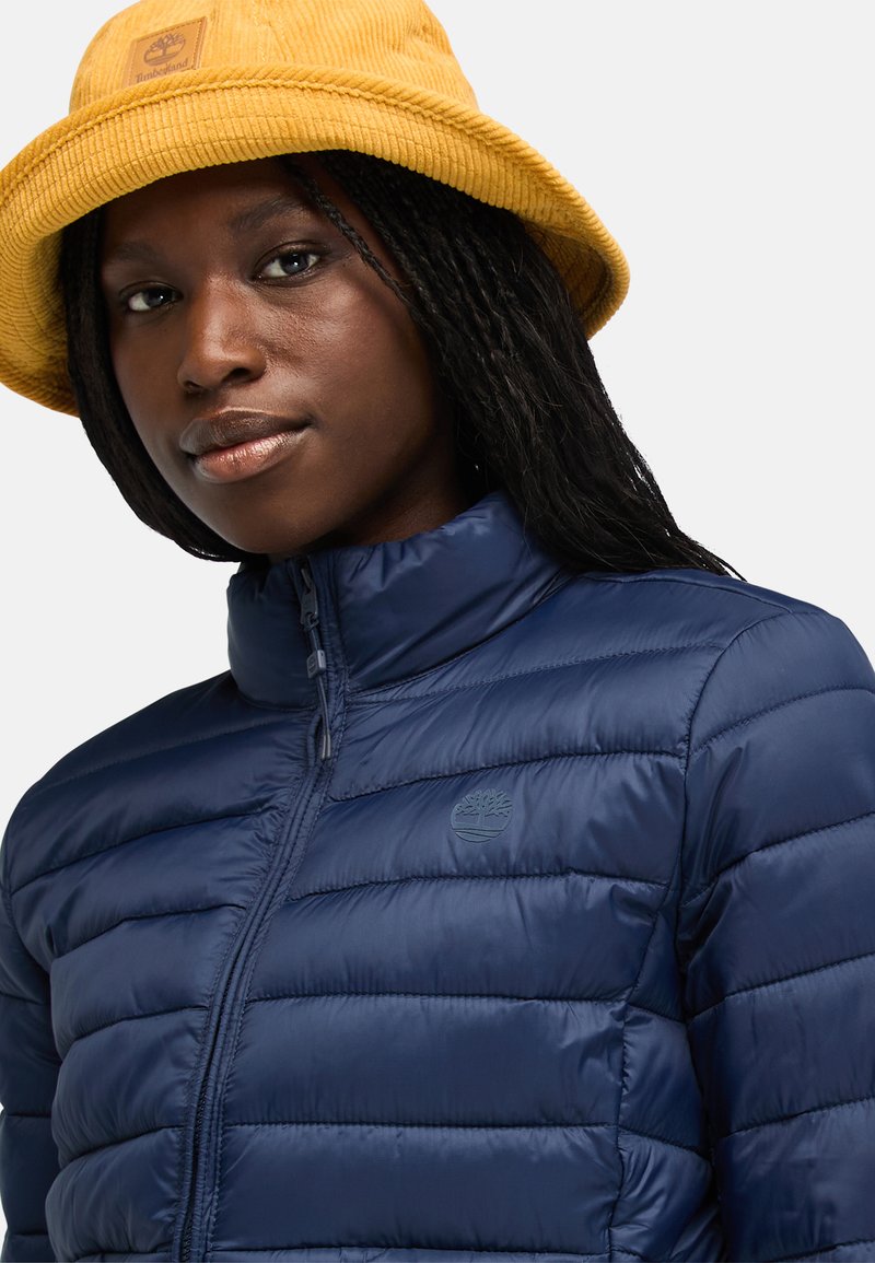 Giacca puffa blu marino con design trapuntato, collo alto e logo Timberland. Abbinata a un cappello a secchiello in corduroy giallo.