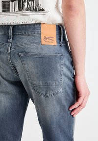 Light blue denim jeans with a leather back patch featuring scissors and “DENHAM.” Fits snugly with a classic five-pocket design and textured fabric.