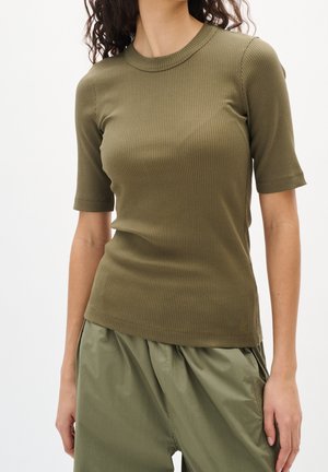 Kobieta ubrana w oliwkowo zielony prążkowany krótki top z krótkim rękawem oraz luźne oliwkowo zielone spodnie, stojąca na jednolitym tle.