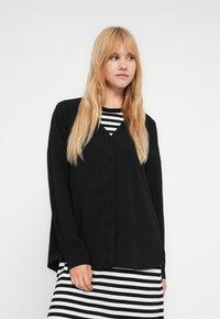 Haut noir à boutons avec une texture douce, manches longues et col en V. Porté par-dessus une robe rayée noir et blanc. Design simple et décontracté.