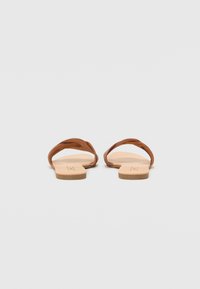 Bruine leren sandalen met een ontwerp van twee gedraaide banden en een platte, tan kleurige zool. Gladde textuur, minimalistische stijl en open achterkant voor gemakkelijk aan- en uittrekken.