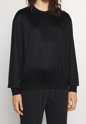 Mujer con una sudadera negra de manga larga holgada y pantalones negros, de pie frente a un fondo claro y liso.