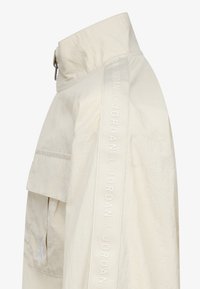 Veste beige clair avec un col haut, dotée d'une grande poche frontale et du texte "JORDAN" le long de la manche, fabriquée en matériau texturé.