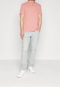 Ljusrosa pikétröja med tre knappar, kombinerad med ljusblå jeans. Modellen bär vita sneakers. Enkel, avslappnad design och passform.